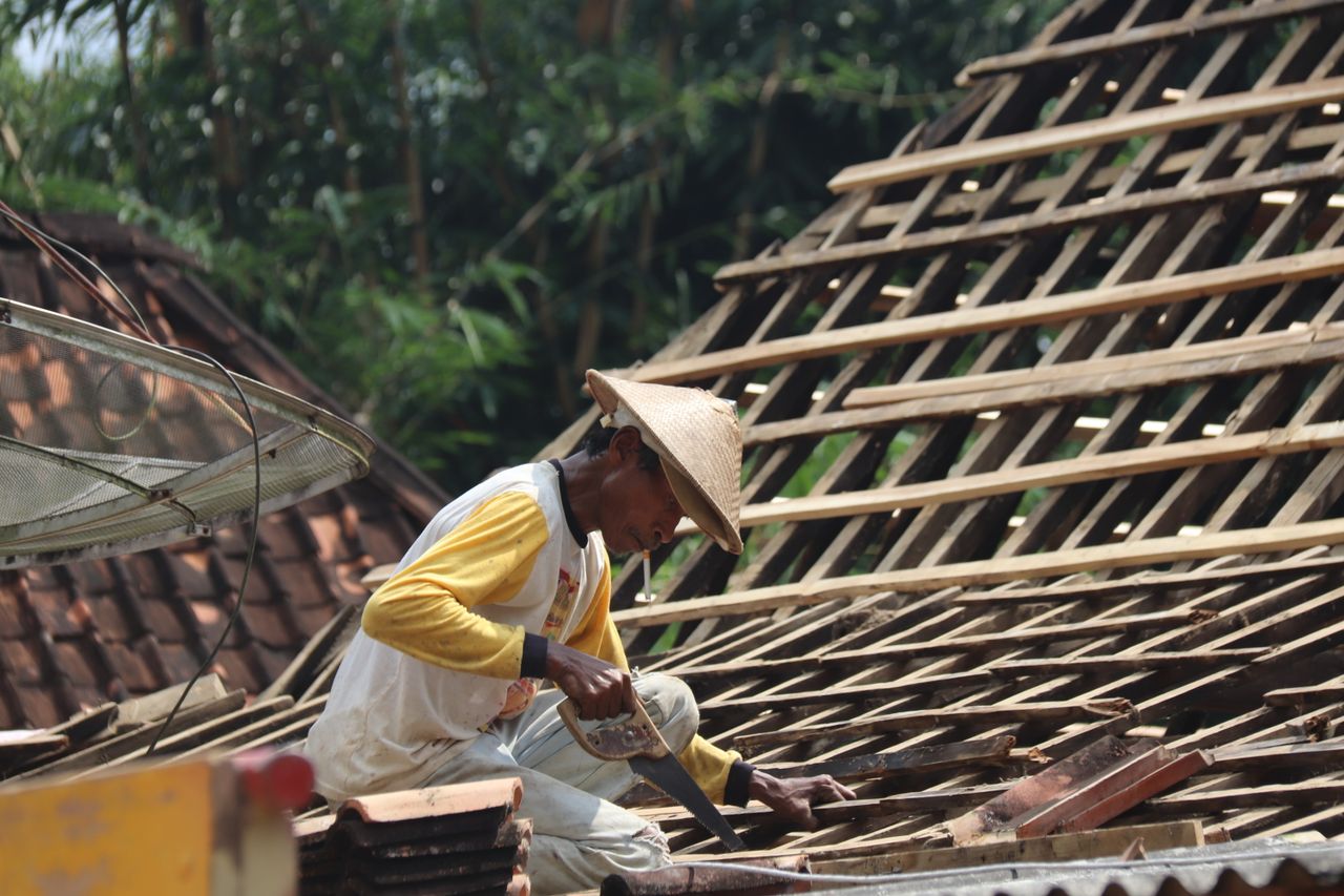 Carpenter working on a wooden roof structure outdoors in a rural setting with traditional tools.